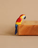 The Bumbu Wooden Amazonian Parrot, a small and colorful handmade figurine featuring red, yellow, and blue feathers, is perched on the edge of a wooden block against a beige background. This eco-friendly toy is crafted from wood and designed for Montessori educational play, appearing to be part of a larger, smooth wooden structure.