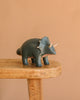 The Konges Sloejd Money Bank - Dino, a small dark green triceratops toy with cream-colored horns, stands on a wooden stool against a plain beige background.
