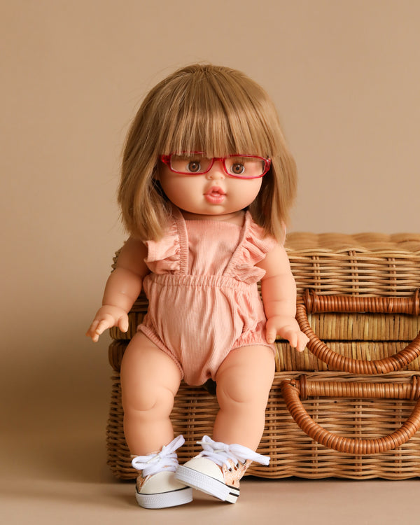 The Minikane Baby Doll (13") - Zoe, featuring short blonde hair and red-framed glasses, is dressed in a stylish peach romper. Posed in front of a woven basket with white sneakers that enhance the neutral beige backdrop, this anatomically correct doll also boasts a natural vanilla scent for added charm.