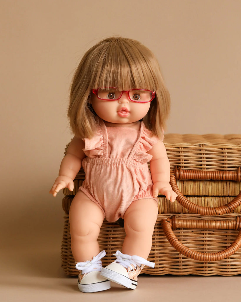 A Minikane doll featuring light brown hair and red glasses poses in front of a woven basket. Designed in France, the doll is dressed in the Lou Retro Romper made from salmon corduroy, complemented by ruffled sleeves and white sneakers. The background is a neutral shade, creating a charming scene.