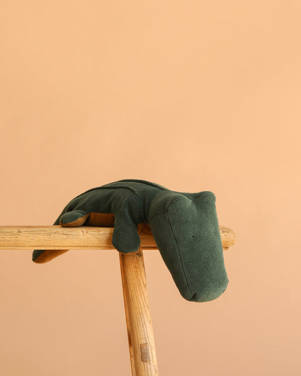 The Maileg Crocodile, Big - Forest Green (ships in approximately one week) is a soft plush toy displayed on a wooden stool with a peach background—ideal as a huggable companion for any plush toy enthusiast.