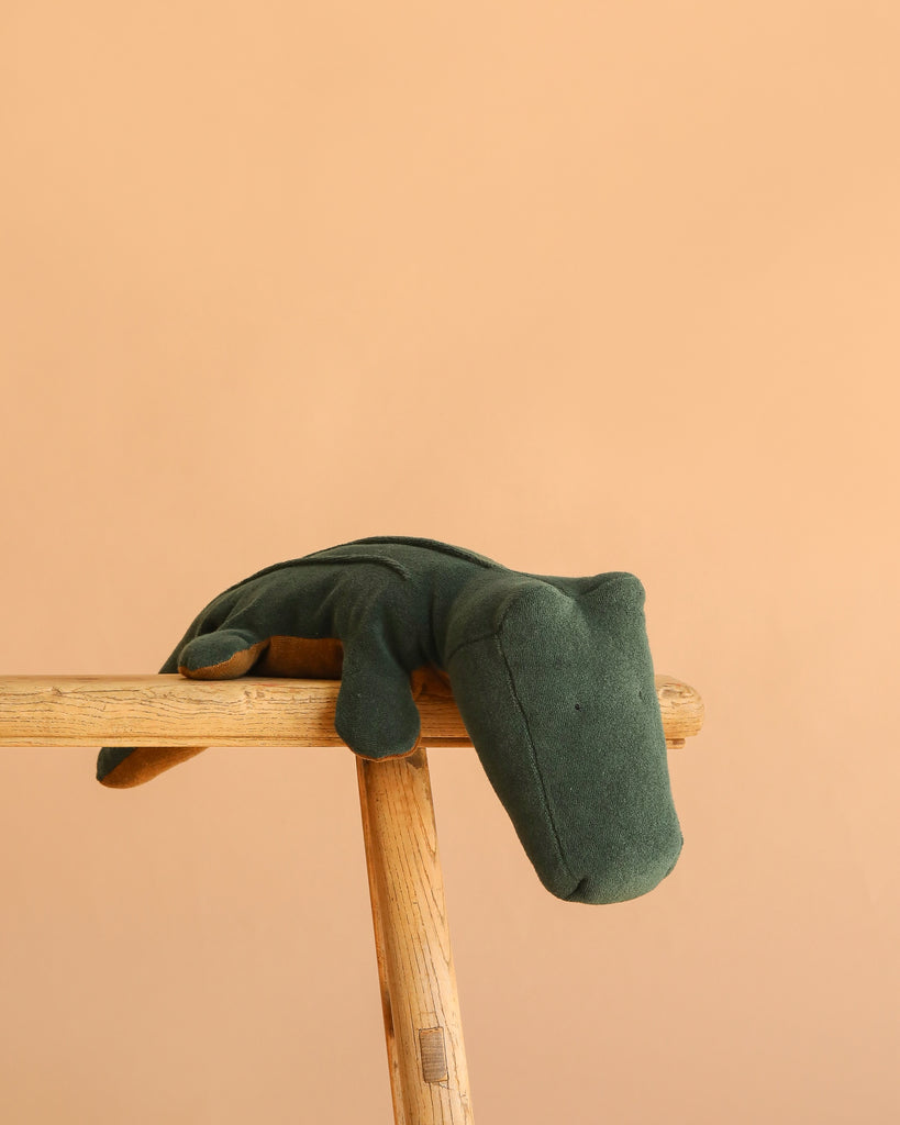 The Maileg Crocodile, Big - Forest Green (ships in approximately one week) is a soft plush toy displayed on a wooden stool with a peach background—ideal as a huggable companion for any plush toy enthusiast.
