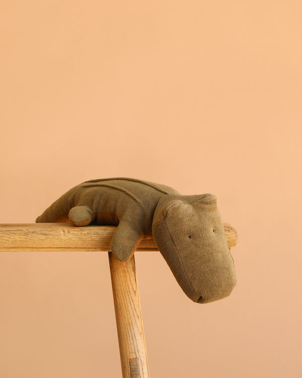 The Maileg Crocodile, Medium - Olive Green is a soft, huggable plush toy shown resting on a light wooden stool against a plain peach-colored background.