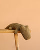The Maileg Crocodile, Medium - Olive Green is a soft, huggable plush toy shown resting on a light wooden stool against a plain peach-colored background.