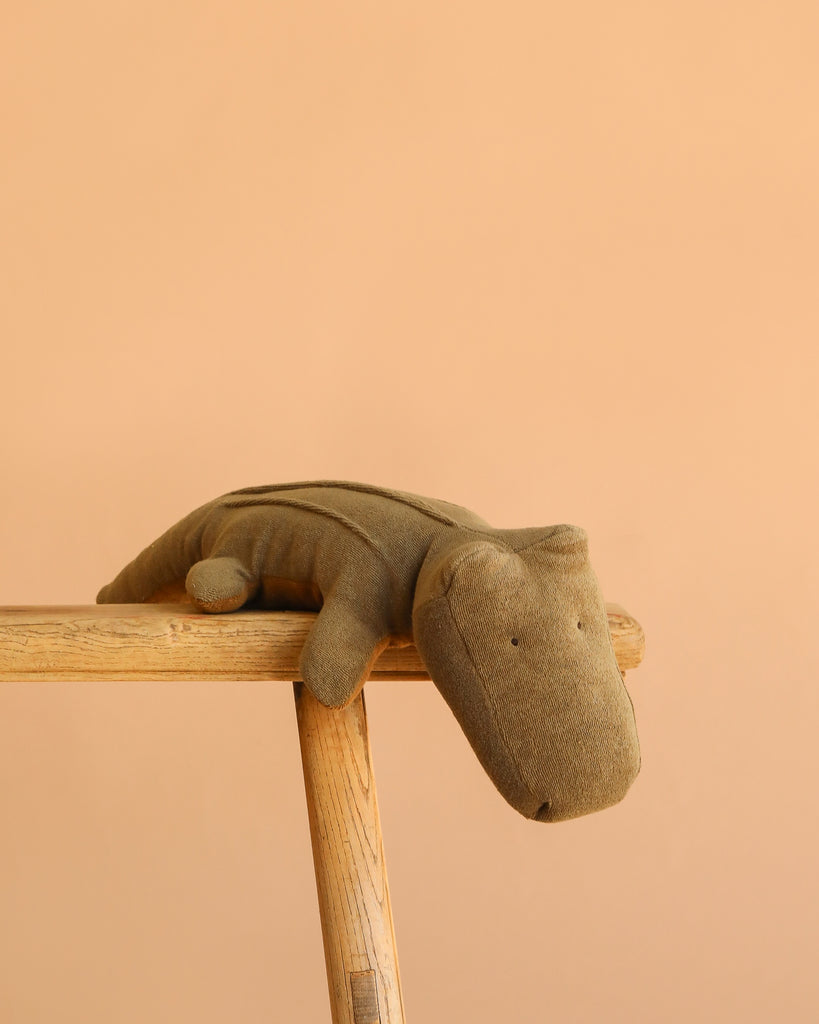The Maileg Crocodile, Medium - Olive Green is a soft, huggable plush toy shown resting on a light wooden stool against a plain peach-colored background.
