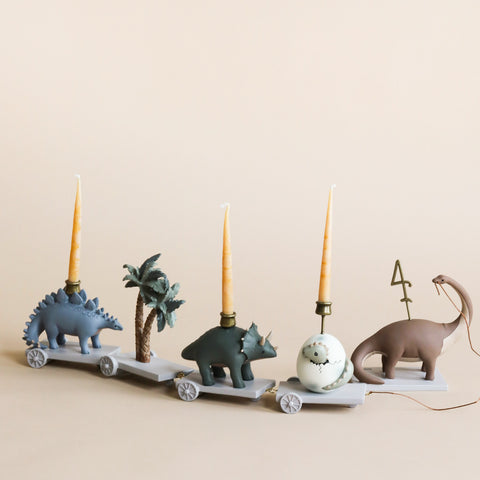Four whimsical candle holders shaped like animals—a Dinosaur Birthday Train With Beeswax Candles, a rhinoceros, an elephant, and a camel—aligned on a light background, each with a tall beige candle in a dinosaur theme
