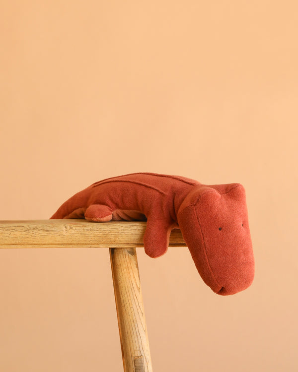 The Maileg Crocodile, Medium - Vintage Rose, a rust-colored plush toy, lounges on the edge of a light wooden bench against a plain peach background.