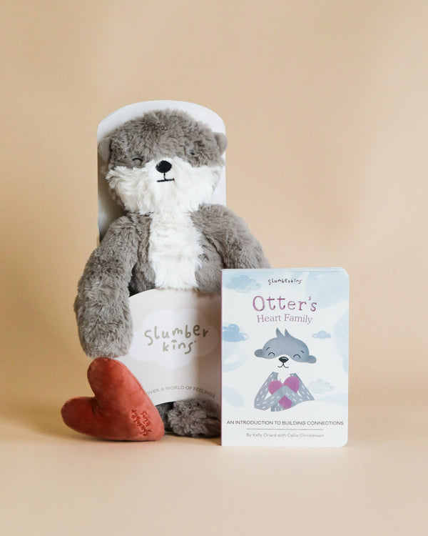 A Slumberkins Otter Kin + Lesson Book - Family Bonding stuffed animal made with hypoallergenic fiberfill and wearing a white bib, sits beside a book titled "Otter's Heart Family" against a soft beige background.