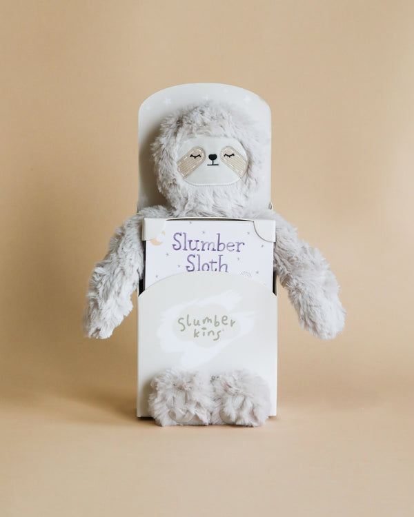 A plush sloth toy with a serene expression, partially inside a white box labeled "Slumberkins Sloth Kin + Lesson Book - Routines" by "slumberkins," designed to support children's routines, set against a soft beige