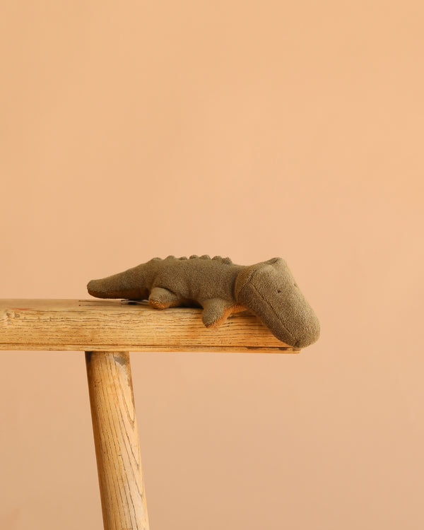 The Maileg Crocodile, Mini - Olive Green rests on the edge of a light wooden table against a plain beige background, perfect for fans of huggable stuffed animals.