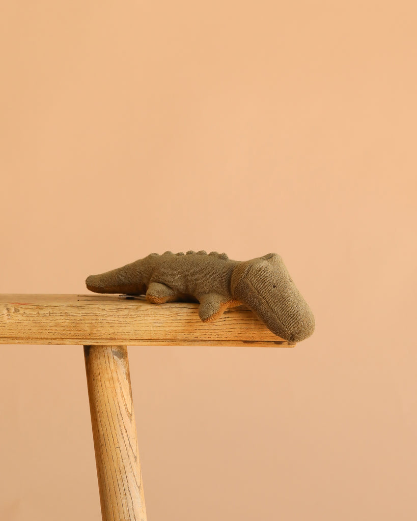 The Maileg Crocodile, Mini - Olive Green rests on the edge of a light wooden table against a plain beige background, perfect for fans of huggable stuffed animals.