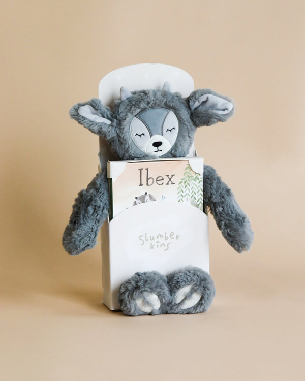 A plush toy resembling a sleepy gray bear, designed as a children's companion, packaged in a box labeled "Slumberkins Ibex Kin + Lesson Book On Emotional Courage" against a soft beige background.