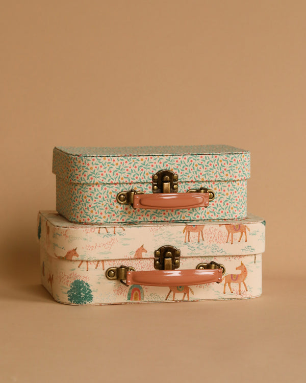 The Maileg 2 Piece Suitcase Set is adorned in pastel colors and consists of two vintage-style suitcases stacked together. The top suitcase, made from Maileg fabrics, showcases a charming floral pattern, while the larger bottom suitcase features a whimsical design with animals and nature scenes. Both suitcases boast sturdy handles and brass latches, making them perfect for stylish storage against a beige backdrop.