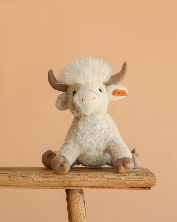The Steiff Harper Highland Cow, featuring cream-colored fur, brown horns, and hooves, sits on a wooden bench against a plain beige background—a charming nursery plush perfect for cuddles.