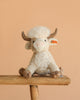 The Steiff Harper Highland Cow, featuring cream-colored fur, brown horns, and hooves, sits on a wooden bench against a plain beige background—a charming nursery plush perfect for cuddles.
