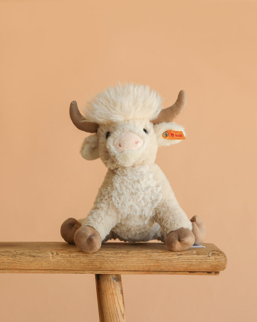 The Steiff Harper Highland Cow, featuring cream-colored fur, brown horns, and hooves, sits on a wooden bench against a plain beige background—a charming nursery plush perfect for cuddles.