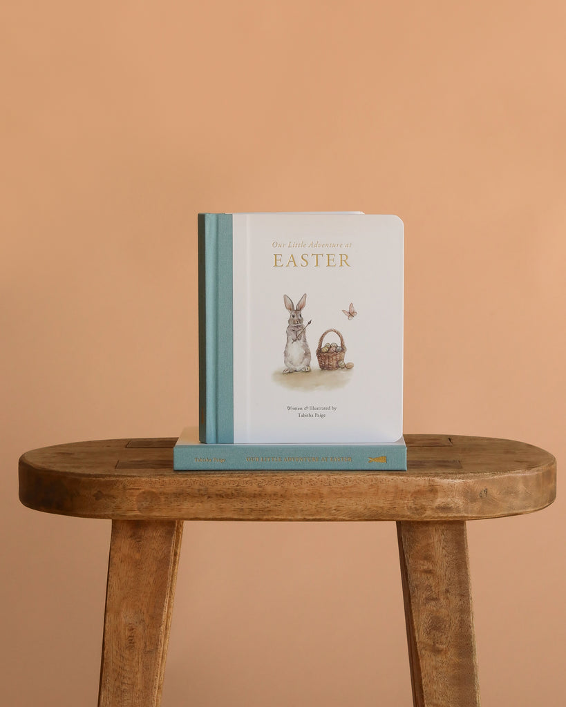 The "Maileg Easter Basket Set," featuring a bunny, handmade basket, and butterfly, is displayed upright on a wooden stool with another item beneath it, set against a peach-colored background.
