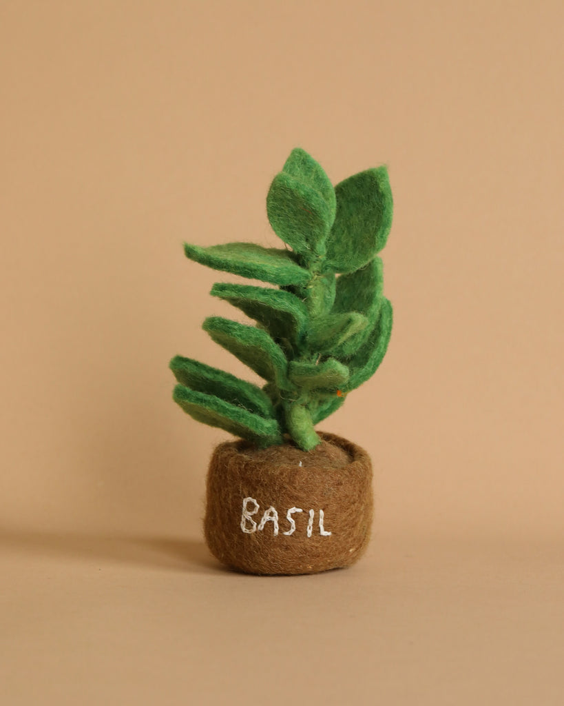 A Handmade Felt Herb Pot featuring a small basil plant with green leaves and a brown base crafted from sustainable materials. The word "BASIL" is written in white on the base, perfect for a pretend kitchen. The beige background beautifully highlights the intricate details of these felted herbs.