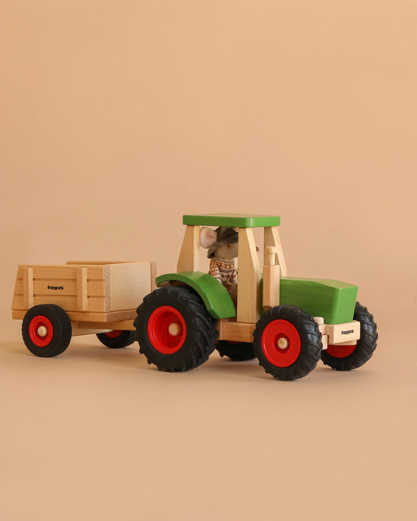 The Maileg x Fagus Limited Edition Tractor Set, featuring a green wooden tractor with red wheels and a matching trailer, rests on a beige surface against a plain beige background.