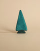 A teal, handcrafted Ostheimer Spruce Tree figurine stands against a light beige background, showcasing a simple and minimalist design.