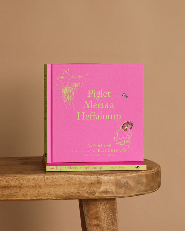 A pink hardcover of "Winnie-the-Pooh: Piglet Meets A Heffalump" sits upright on a wooden stool with another copy flat underneath, against a plain beige background—ideal for fans of classic bedtime stories and Winnie the Pooh.