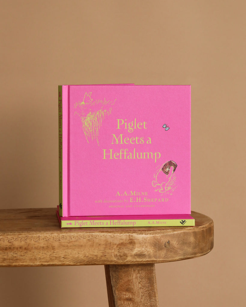 A pink hardcover of "Winnie-the-Pooh: Piglet Meets A Heffalump" sits upright on a wooden stool with another copy flat underneath, against a plain beige background—ideal for fans of classic bedtime stories and Winnie the Pooh.