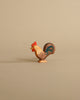 A small, painted rooster from the Ostheimer Handmade Wooden Farm Animals Set, featuring a red comb, brown body, and blue tail, stands on a light beige background—a charming handcrafted toy.