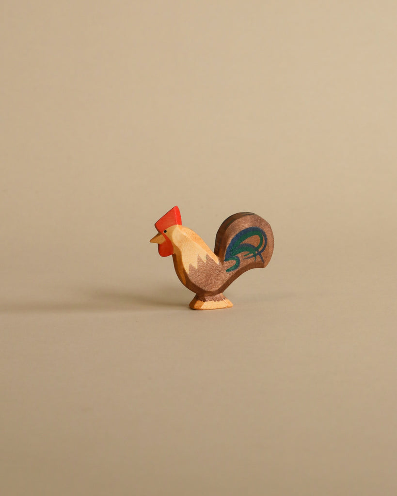 A small, painted rooster from the Ostheimer Handmade Wooden Farm Animals Set, featuring a red comb, brown body, and blue tail, stands on a light beige background—a charming handcrafted toy.