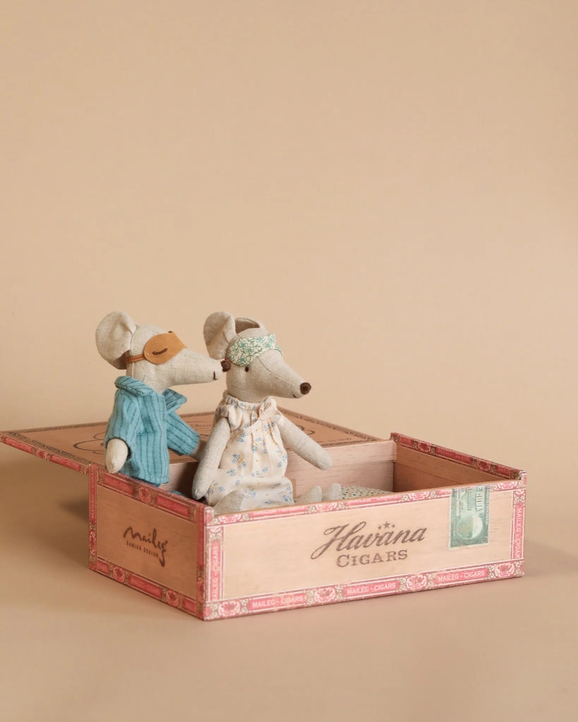 Two stuffed toy mice from the Maileg Christmas Set sit in an open wooden cigar box. One mouse is dressed in a blue outfit with a sleep mask, and the other wears a floral dress with a headband. The box, labeled "Havina Cigars" and decorated with stamps, poses a small parts choking hazard for children under three against the beige background.