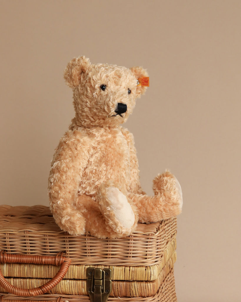 A Steiff Elmar Teddy Bear, 12 inches in light brown with a fluffy texture, black nose, and eyes sits against a beige background on top of a wicker basket with a metal clasp. Crafted from premium-grade materials, the handmade bear's posture is upright with its legs extended forward.