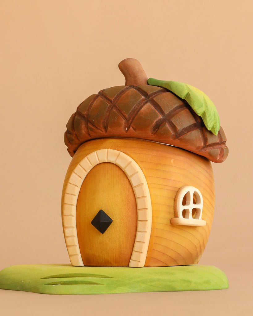 The Bumbu Handmade Wooden Acorn House features a whimsical acorn shape with a wooden door, small window, and leaf roof, sitting on a green patch against a beige background.