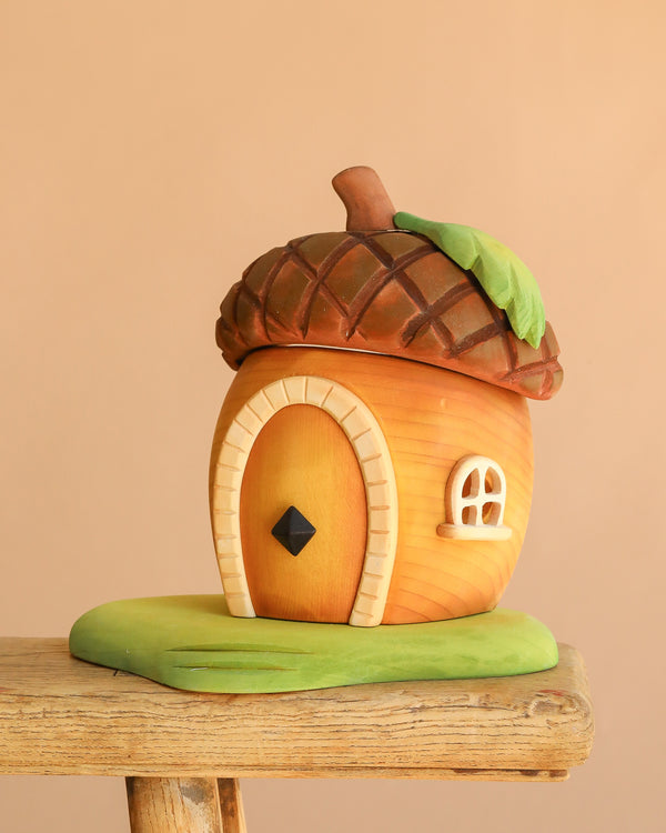 The Bumbu Handmade Wooden Acorn House is a whimsical toy with a rounded wooden door, tiny window, and green base, perfect as a fairy home. It sits on a wood surface with a plain beige background.