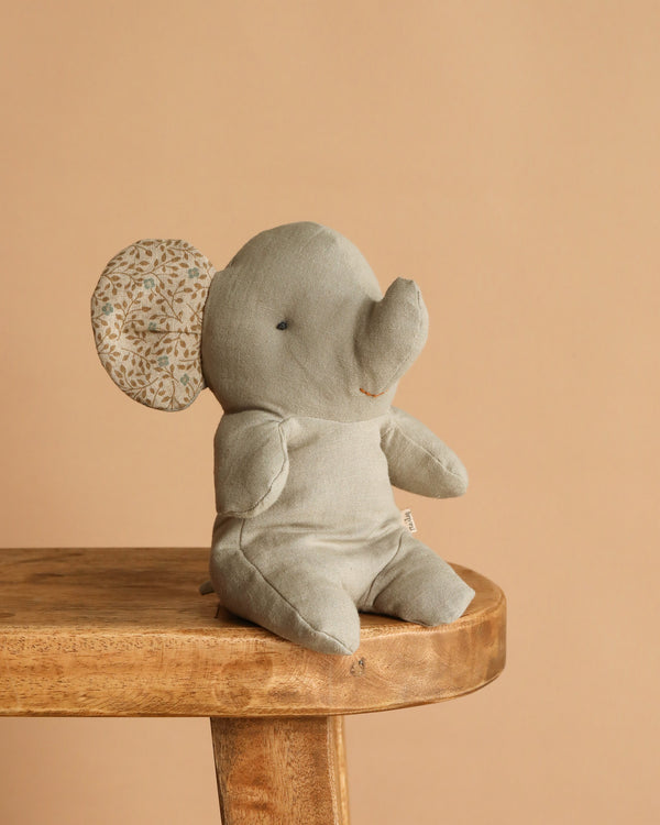 The Maileg Elephant, Small - Iron Grey, with soft plush and patterned ears, sits charmingly on a wooden bench against a beige background—a cute addition to any Safari friends collection.