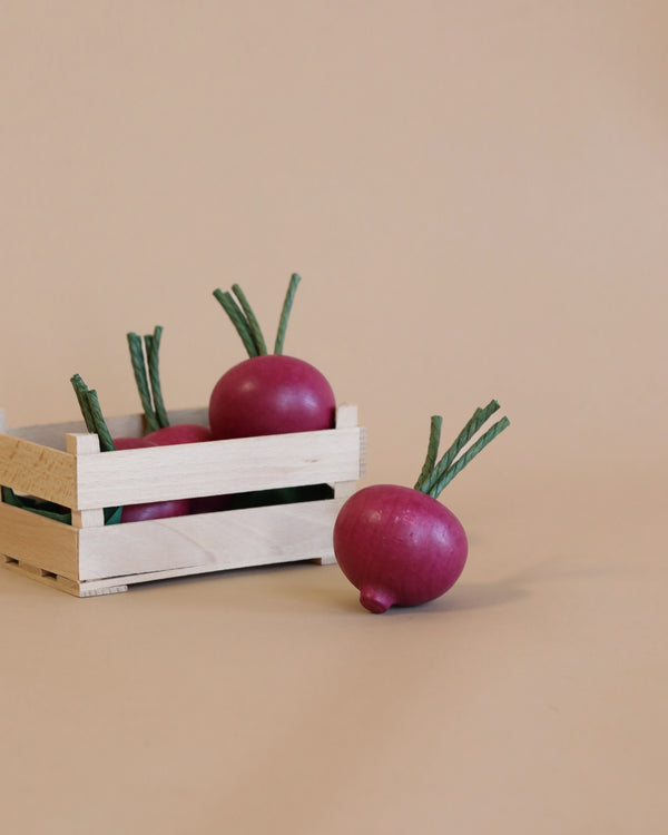 Three purple radishes with green stems, one outside and two inside a small wooden crate, against a light beige background. This realistic Erzi Beetroot Pretend Food is handcrafted in Germany.