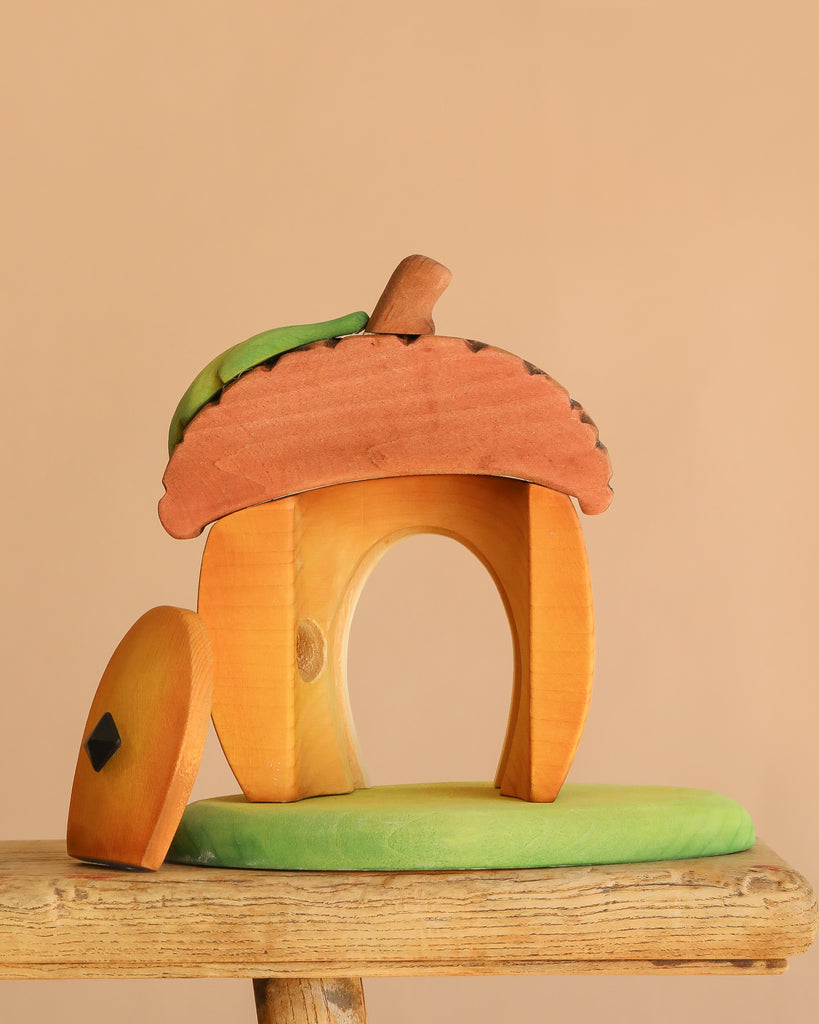 The Bumbu Handmade Wooden Acorn House, shaped like an acorn with a brown textured roof, green leaf detail, and open door, sits on a green base atop a wooden surface against a beige background.