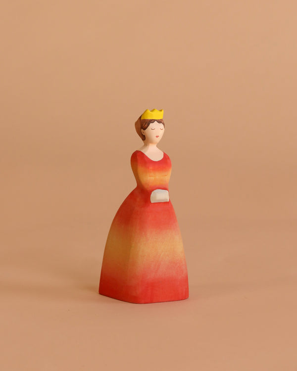 The Bumbu Handmade Wooden Princess features a woman in a yellow crown and red-orange dress with hands folded, ideal for Waldorf-inspired play, shown against a beige background.