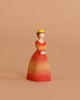 The Bumbu Handmade Wooden Princess features a woman in a yellow crown and red-orange dress with hands folded, ideal for Waldorf-inspired play, shown against a beige background.