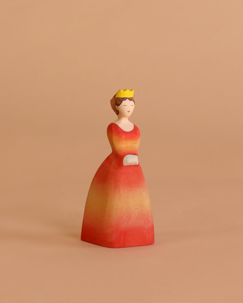 The Bumbu Handmade Wooden Princess features a woman in a yellow crown and red-orange dress with hands folded, ideal for Waldorf-inspired play, shown against a beige background.