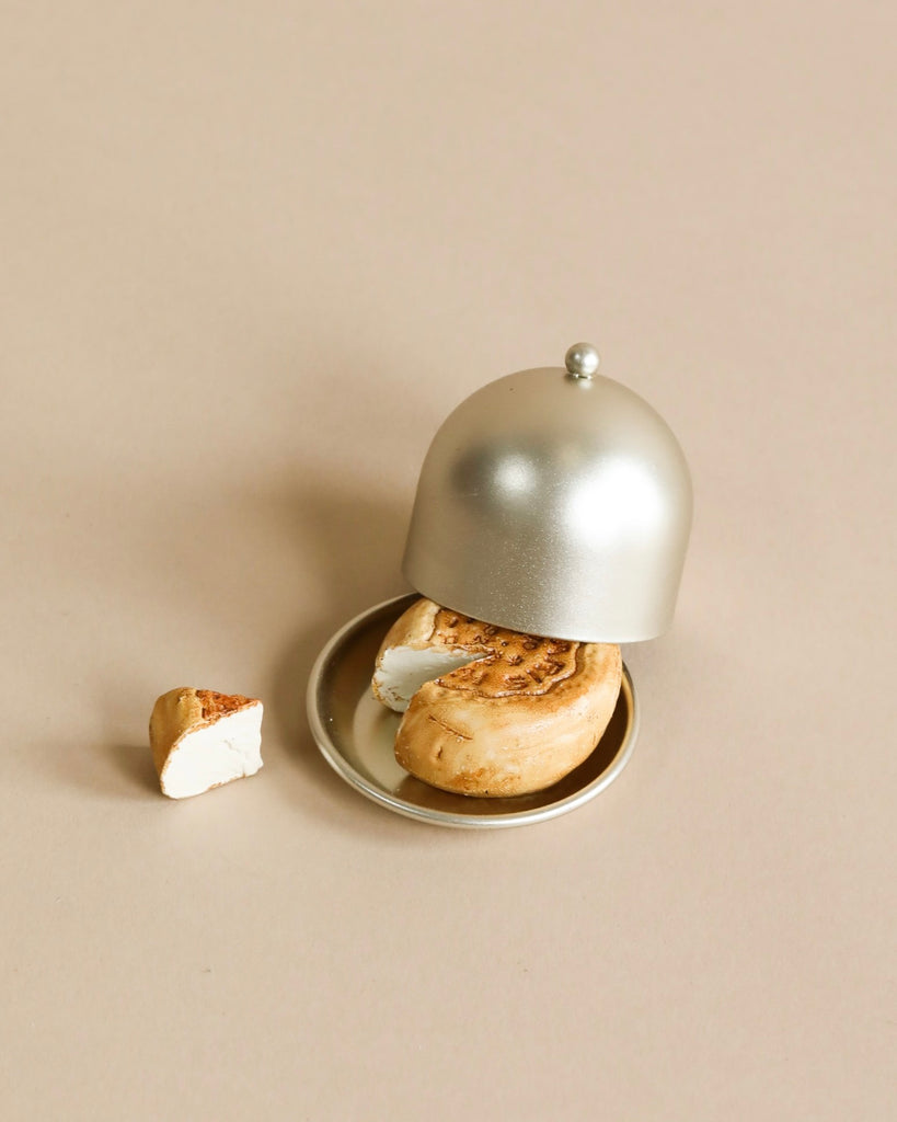 A cinnamon roll with a bite missing sits on a small plate, partially covered by a silver cloche—perfectly sized for the Maileg Grand Dining Room Set or any dollhouse display. A separate piece of the roll rests beside the plate on a beige surface.