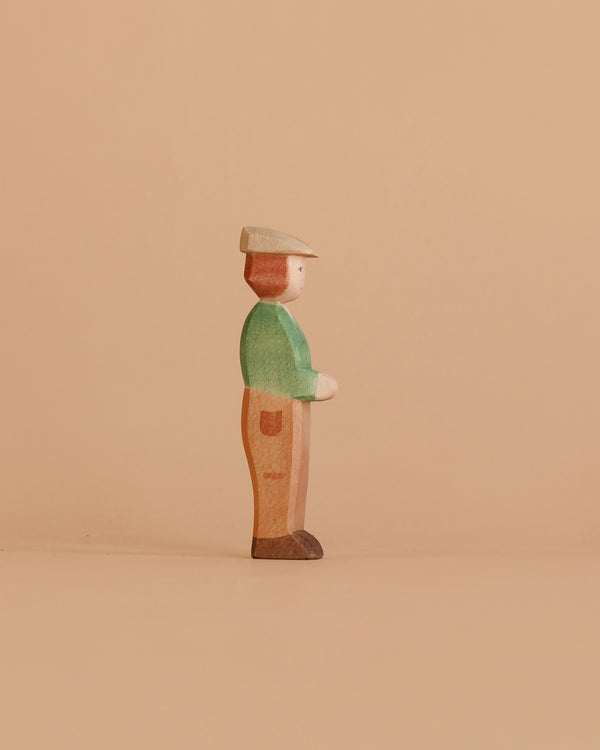 The Ostheimer Young Farmer, a handcrafted wooden figurine in a green shirt, brown pants, and beige hat, stands in profile against a plain beige background.