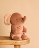 A Maileg Elephant in Carry Cot sits on a wooden stool against a beige background, its large ears featuring a dotted inner pattern as it faces slightly right.