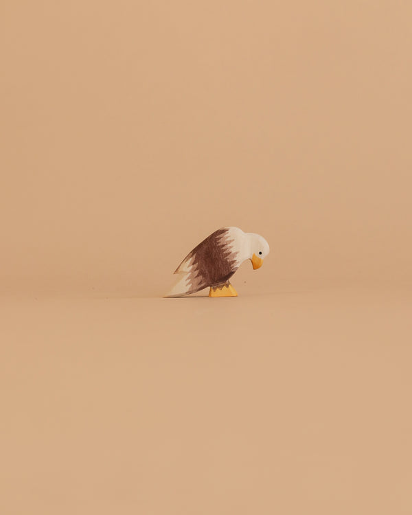 The Ostheimer Bird - Bald Eagle, Sitting features a small wooden eagle with a white head, brown wings, and yellow beak and feet. It is displayed on a plain beige surface against a matching beige background.