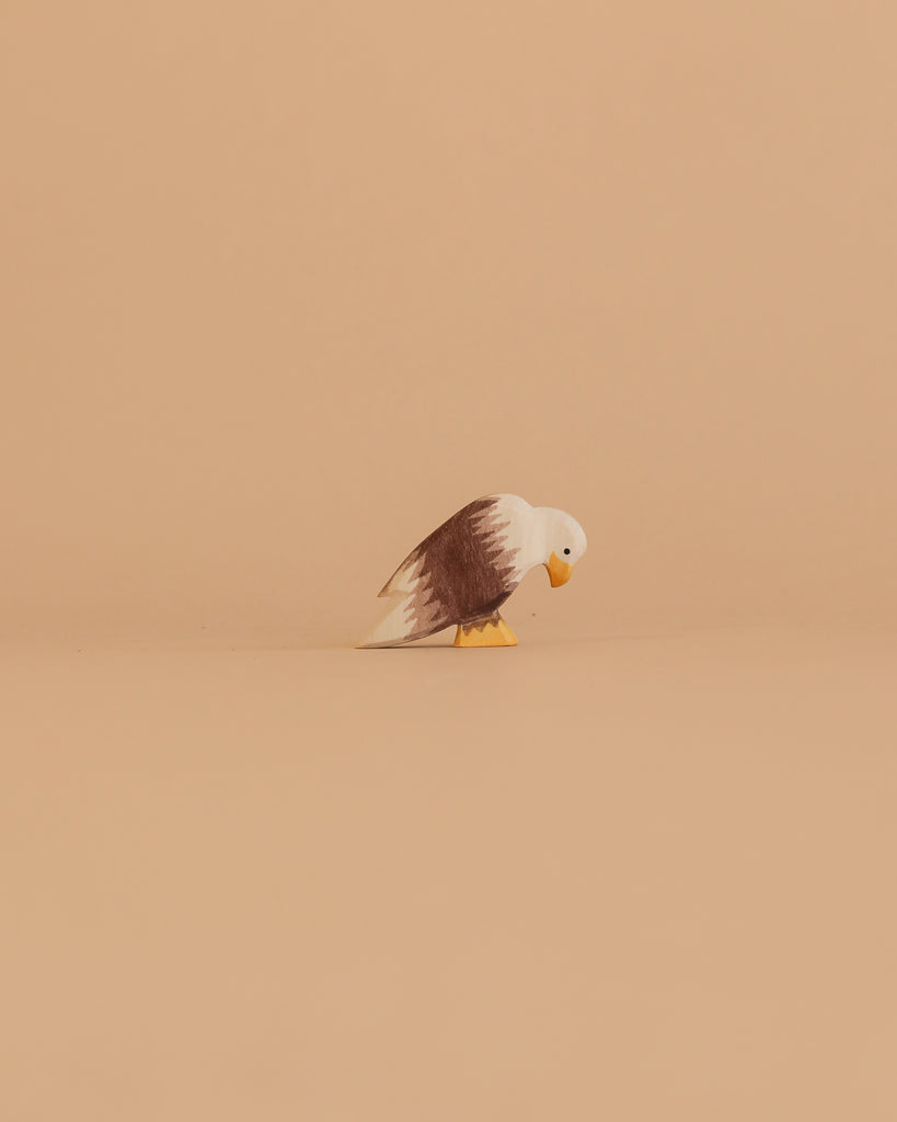 The Ostheimer Bird - Bald Eagle, Sitting features a small wooden eagle with a white head, brown wings, and yellow beak and feet. It is displayed on a plain beige surface against a matching beige background.