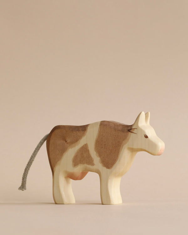 A sustainable wooden Handmade Holzwald Cow with brown and white patches, standing against a light beige background. The cow has a detailed tail and ears, positioned in a stationary stance.