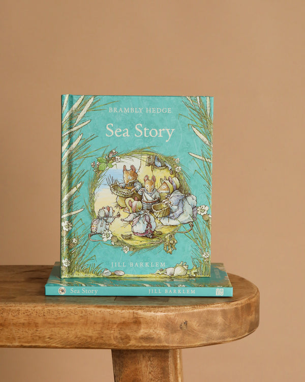 A turquoise hardcover children's book, "Sea Story: Jill Barklem," is displayed upright on a wooden table with another copy lying flat beneath it, set against a plain beige background.