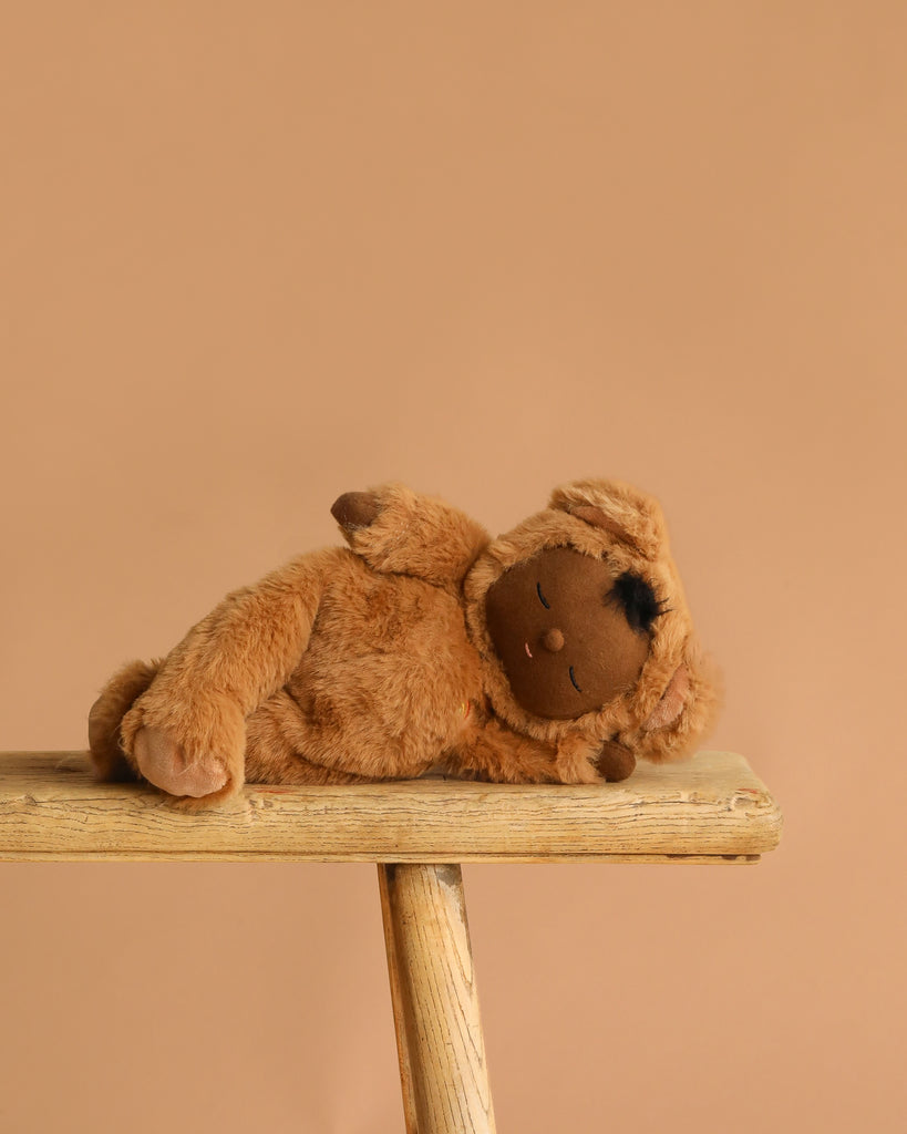 The Olli Ella Cozy Dinkum - Teddy Mini, a plush bear with brown fur and a dark brown face, rests on its side atop a wooden bench against a simple light brown background.