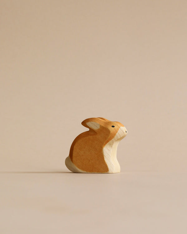 A high-quality Handmade Holzwald Sitting Rabbit with a smooth, curved design and two-tone color stands against a plain, light beige background. The rabbit is positioned to the left, appearing contemplative.