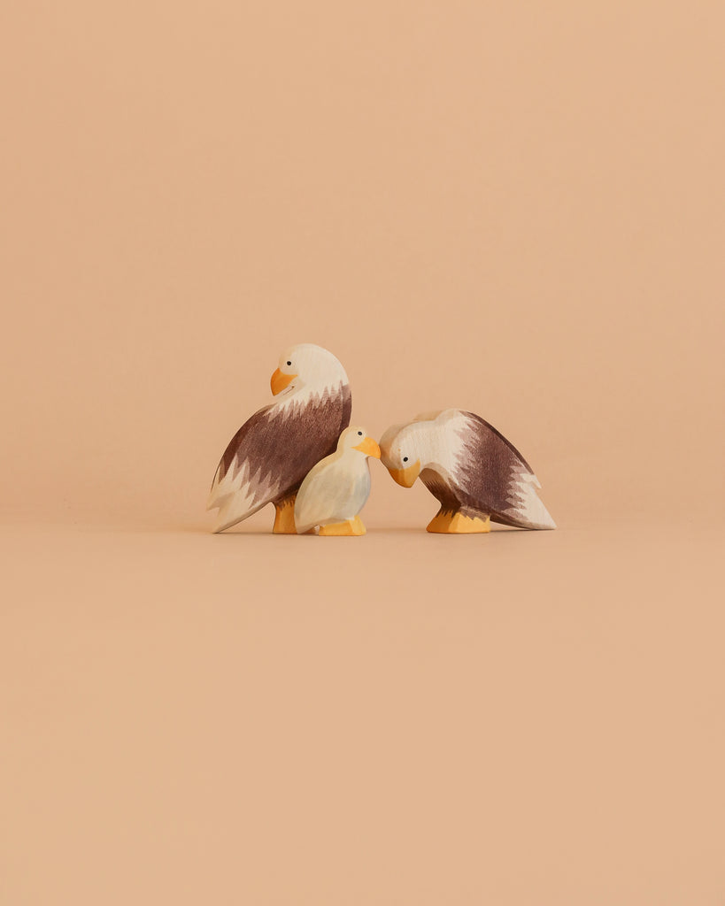 The Ostheimer Bald Eagle Family Set features two large and one small handcrafted figurine, arranged together on a beige surface with a matching background, evoking the charm of classic Ostheimer wooden toys.