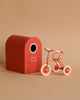The Maileg Big Sister Tricycle Set, featuring a small red and white mouse toy, stands by a red "Abri à Tricycle" box shaped like a birdhouse against a beige background.