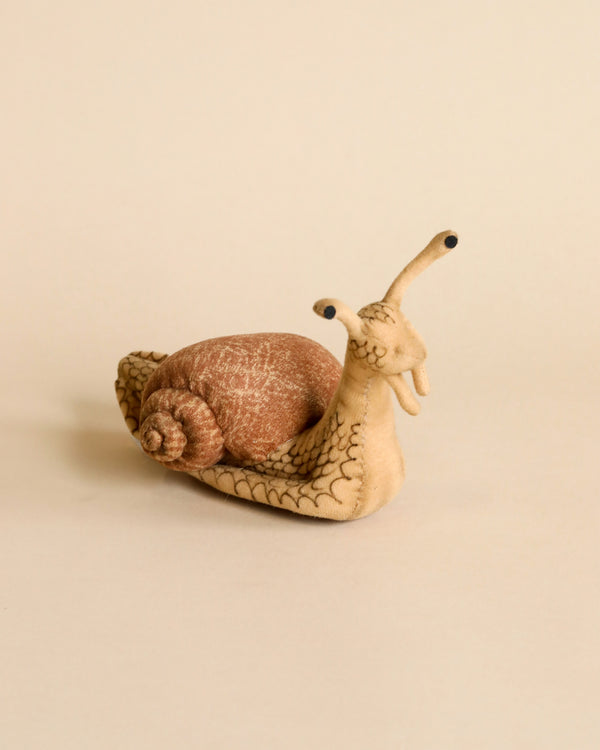 A Snail Stuffed Animal with a brown shell and cream-colored body displayed on a plain beige background. The snail has cute, black eyes on stalks and detailed stitching of hand sewn plush animals.